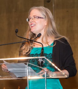 Suzanne Collins standing at a podium speaking to a crowd.