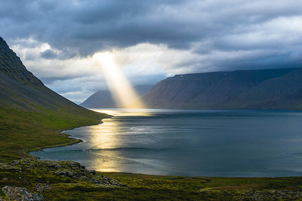 coastal beach with mountains in the distance and a ray of sunlight shining on the ocean from an otherwise cloudy sky