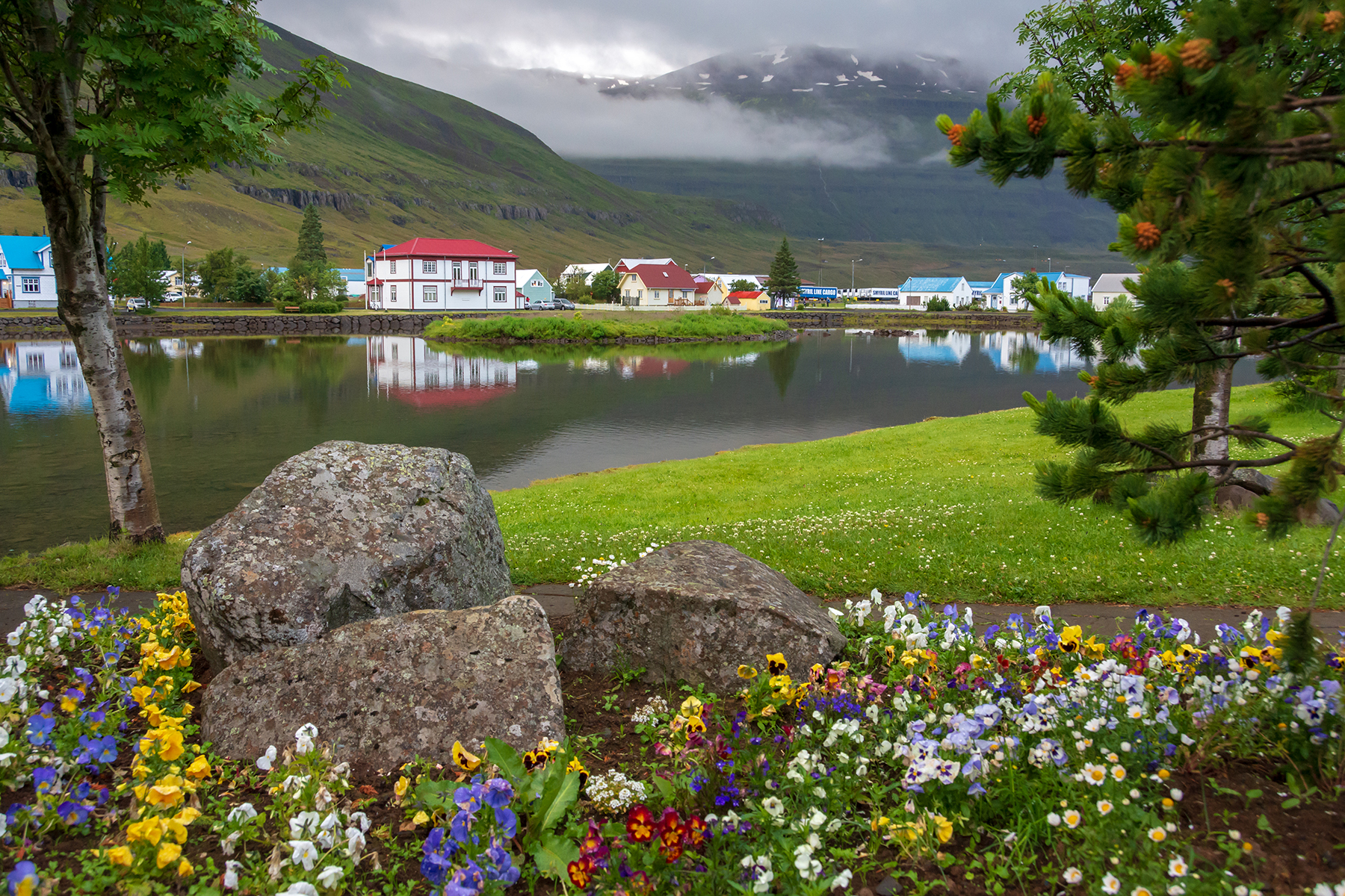 A small town sits across a narrow river with flowers blooming.