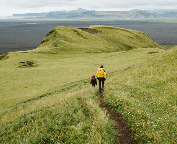 Two people walking down a mountain trail with the ocean in front of them